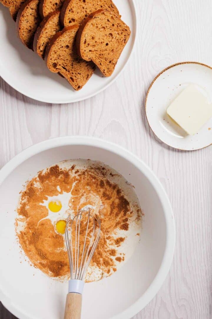 A bowl with gluten-free flour, two egg yolks, and cinnamon sits ready with a whisk. Nearby are a plate of brown bread slices and a small plate holding butter. These items rest on a light wood surface, perfect for creating a delicious gluten-free French toast casserole.