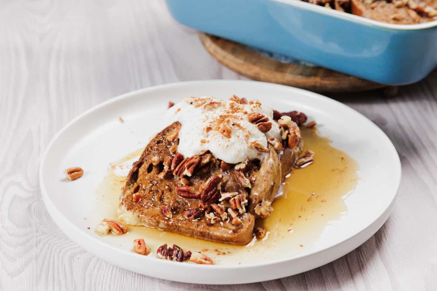 A plate of gluten-free French toast topped with whipped cream, pecans, and caramel sauce rests in a pool of syrup. In the background, a blue baking dish holds more of this delightful casserole on a light-colored wood table.