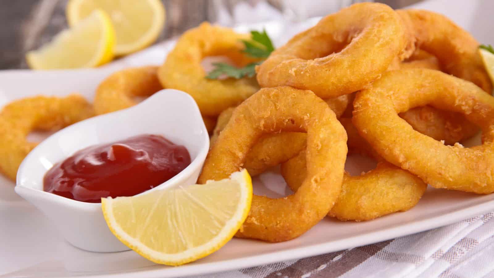 A plate of golden-brown onion rings is served with a small dipping bowl of red sauce, likely ketchup. A lemon wedge is placed beside the onion rings on a white plate. The background includes a lemon slice and some greenery.