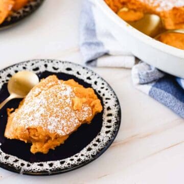 A slice of sweet potato casserole with a dusting of powdered sugar is served on a black and white plate. A gold spoon rests beside it. In the background is a casserole dish with more of the dessert and a white and blue towel.