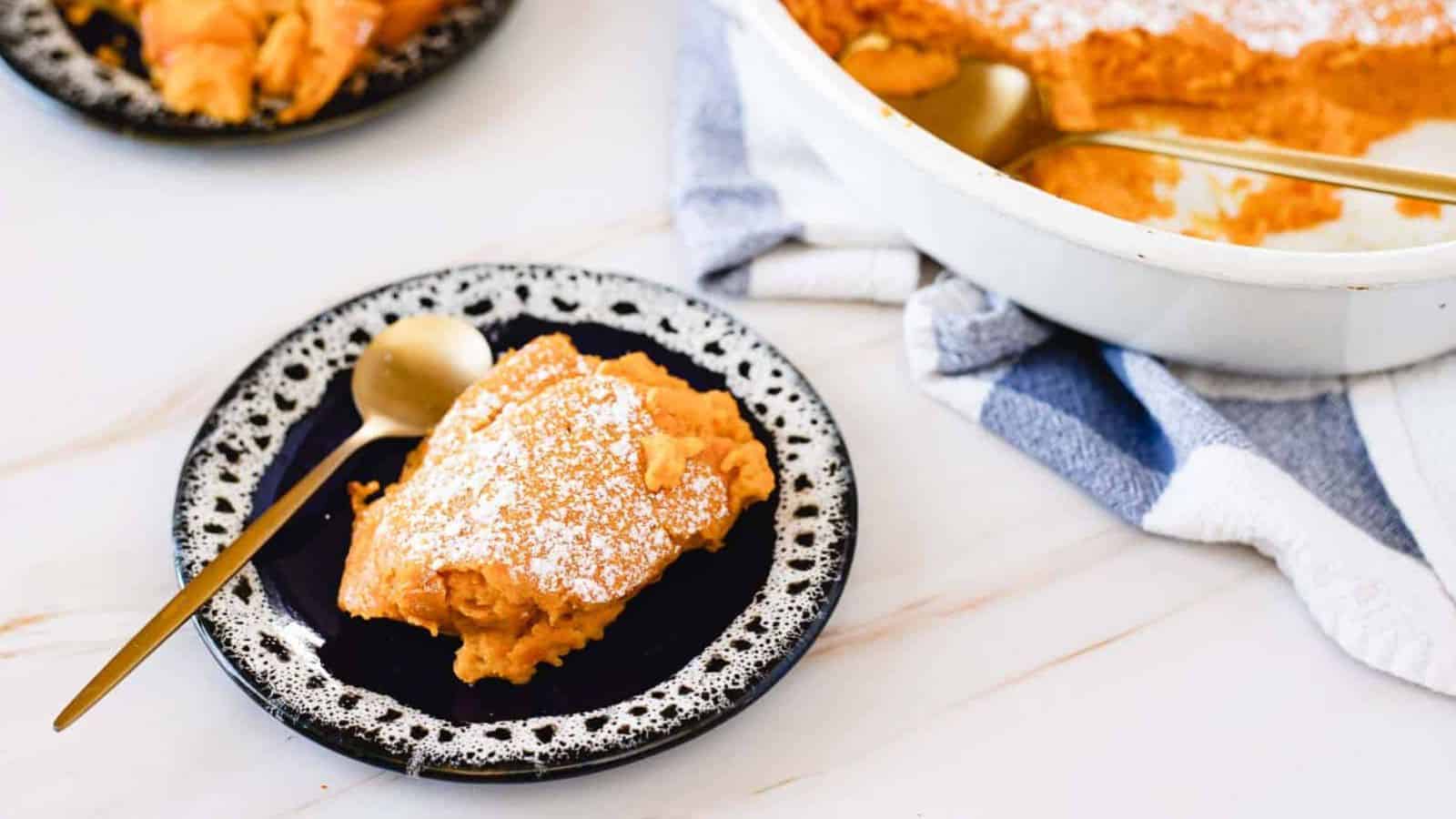 A slice of sweet potato casserole with a dusting of powdered sugar is served on a black and white plate. A gold spoon rests beside it. In the background is a casserole dish with more of the dessert and a white and blue towel.