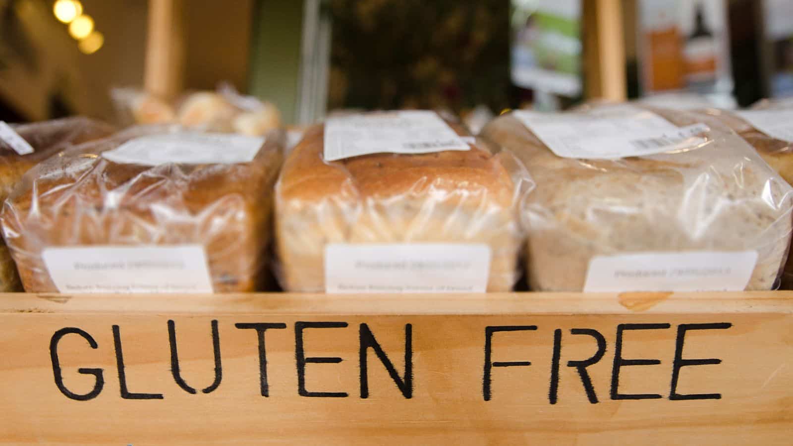 Breads in clear packaging are displayed on a wooden shelf with "GLUTEN FREE" written on the front. Each loaf has a label with product details. The background is slightly blurred, focusing attention on the gluten-free bread selection.