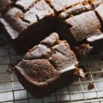 A slice of gluten-free chocolate cake dusted with powdered sugar rests on a cooling rack. The moist cake, resembling a gingerbread loaf with its cracked top and slightly rough texture, sits among other slices, emphasizing a homemade aesthetic.