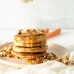 A stack of pancakes reminiscent of carrot cake is topped with chopped walnuts on a plate. Syrup cascades over them, adding sweetness, while two blurred carrots offer a hint of inspiration in the background. Scattered walnuts complete this gluten-free delight set against a light surface.