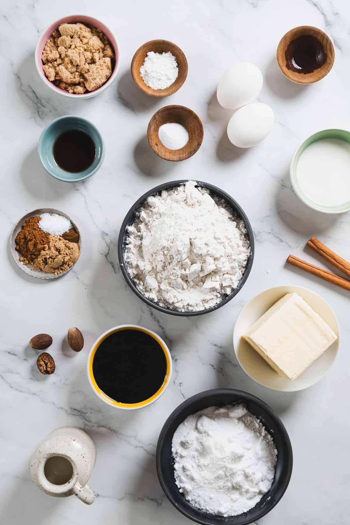 A flat lay of gluten-free baking ingredients on a white marble surface, featuring bowls of brown sugar, flour, and powdered sugar. Eggs, butter, vanilla, molasses for gingerbread loafs, baking soda, salt, nutmeg, cinnamon sticks, and milk are arranged around them.