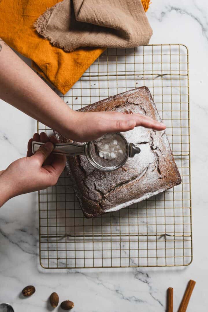 A person sprinkles powdered sugar onto a square chocolate cake using a sifter. The gluten-free cake rests on a cooling rack over a marble surface. Nearby lie a brown cloth, a yellow cloth, and scattered nuts and cinnamon sticks, evoking the cozy scent of gingerbread.