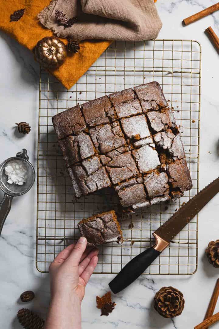 A square gingerbread cake loaf, now cut into pieces, rests invitingly on a cooling rack. A hand reaches for one piece, while nearby are pinecones, cinnamon sticks, and a sieve with sugar on the marble surface. Decorative pumpkins add charm alongside a cloth in the top left corner.