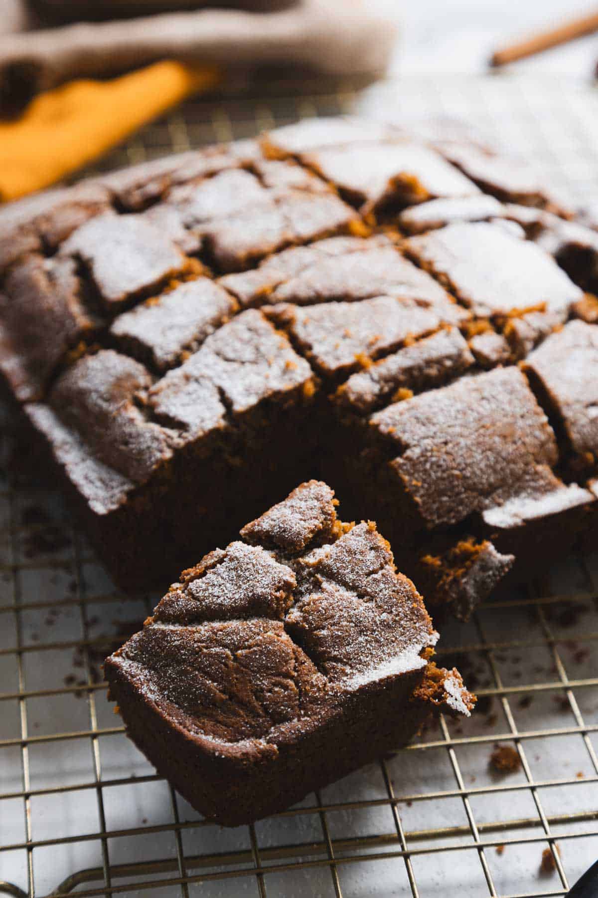 A cooling rack holds a sliced, gluten-free chocolate cake. The cake, divided into squares, has a slightly cracked surface with a dusting of powdered sugar. One piece is pulled out from the grid, resting on the rack.
