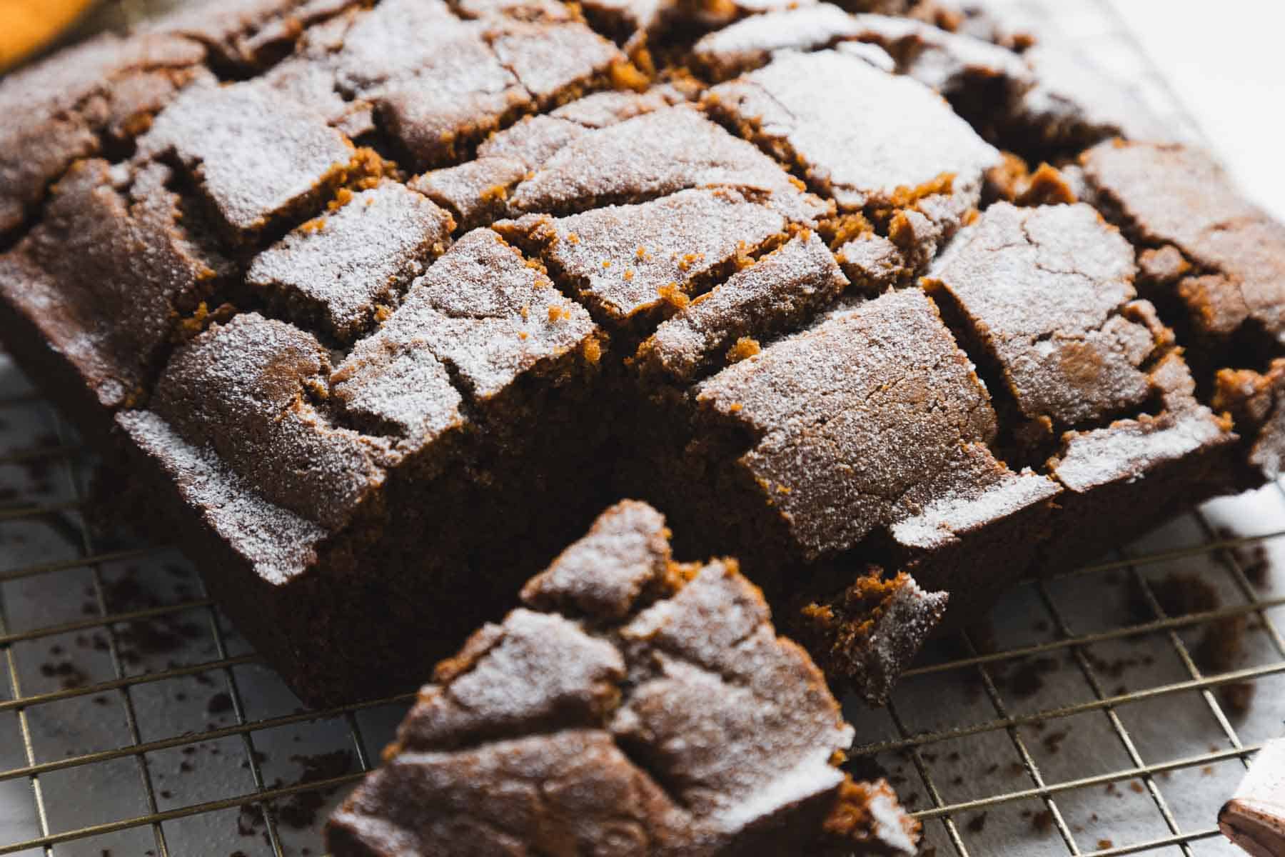 A close-up of sliced gingerbread loaf on a cooling rack. The cake is dusted with powdered sugar, revealing a cracked surface texture. The irregularly shaped slices showcase the loaf's moist interior, making it an irresistible treat.
