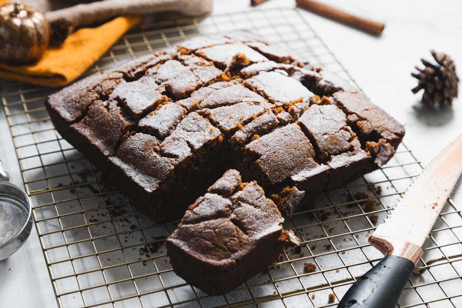A rectangular Gluten-free Gingerbread Loaf, cut into square pieces, sits on a metal cooling rack. A large knife rests nearby, and some cake pieces are missing. Spices and a pinecone add a festive touch in the background.