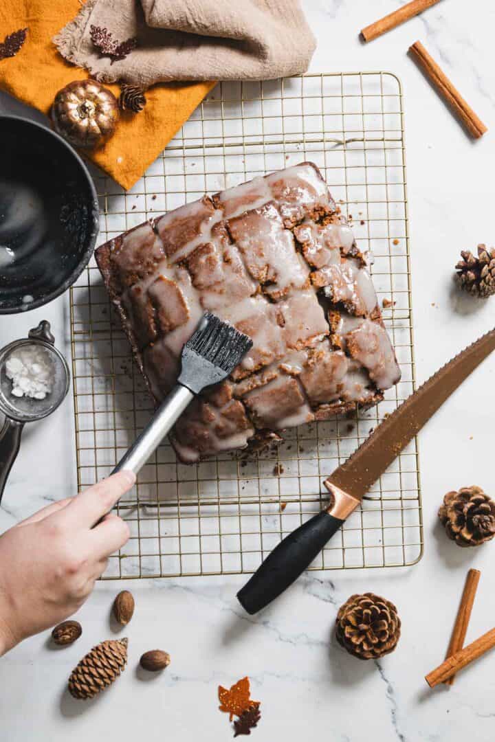 A steady hand brushes glaze onto a square cake on a cooling rack, transforming it into a gingerbread delight. The scene features a knife, small decorative pumpkins, pine cones, cinnamon sticks, and scattered nuts on a marble surface. Nearby, a black bowl and sieve hold powdered sugar for the loaf.