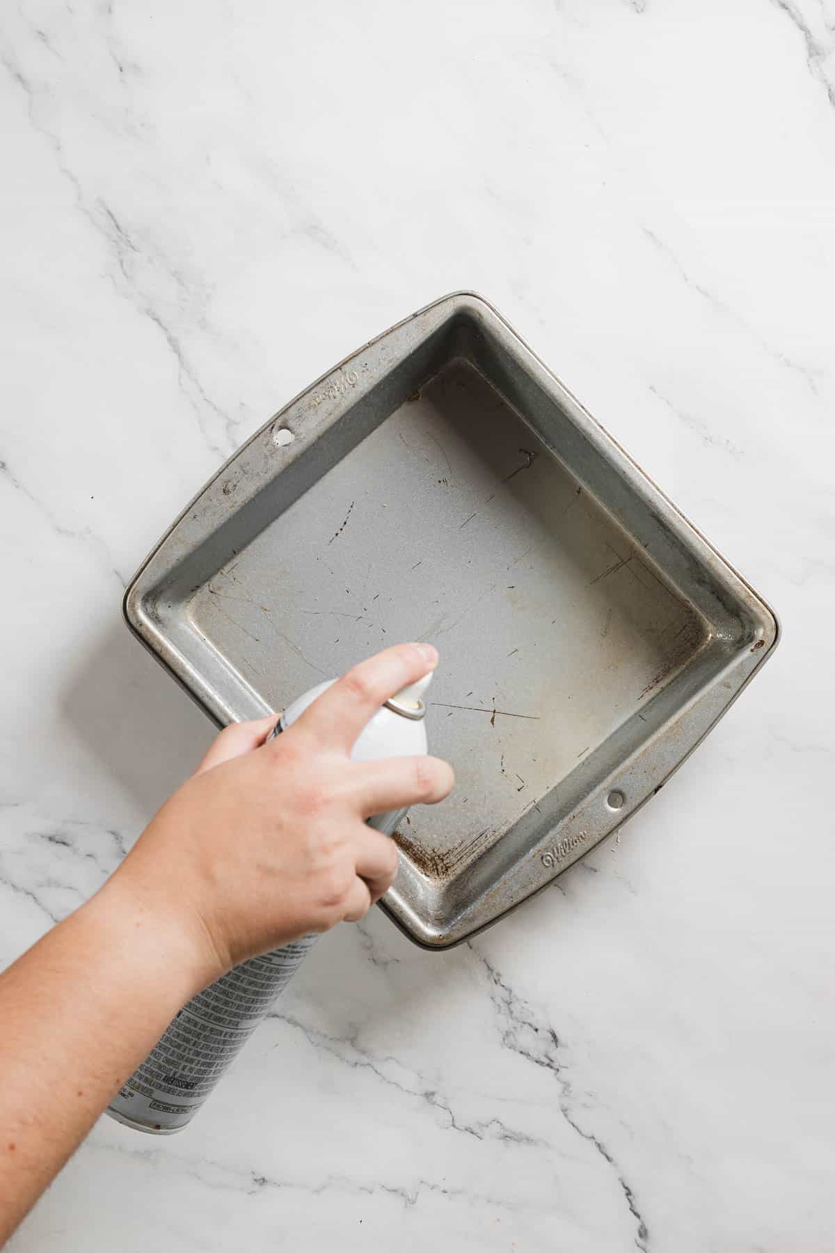 A hand is gently holding a can and spraying a light coating of oil onto a square metal baking pan, ready for a gluten-free creation. The pan rests elegantly on a white marble surface, waiting to bring delicious treats to life.