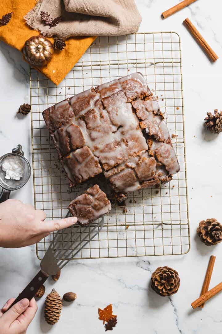 A person cuts a gluten-free square cake glazed with icing on a cooling rack. The surface is sprinkled with small decorative items like pinecones, acorns, and cinnamon sticks. A sifter with powdered sugar rests beside the cake.