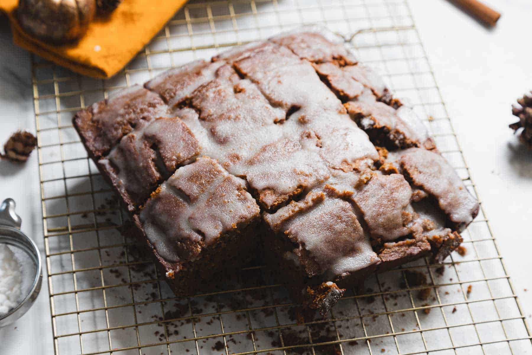 A glazed gingerbread loaf rests on a wire cooling rack, partially cut into squares. It boasts a light icing on top. Surrounding the cake are scattered walnuts, a cinnamon stick, and a small sieve with powdered sugar.