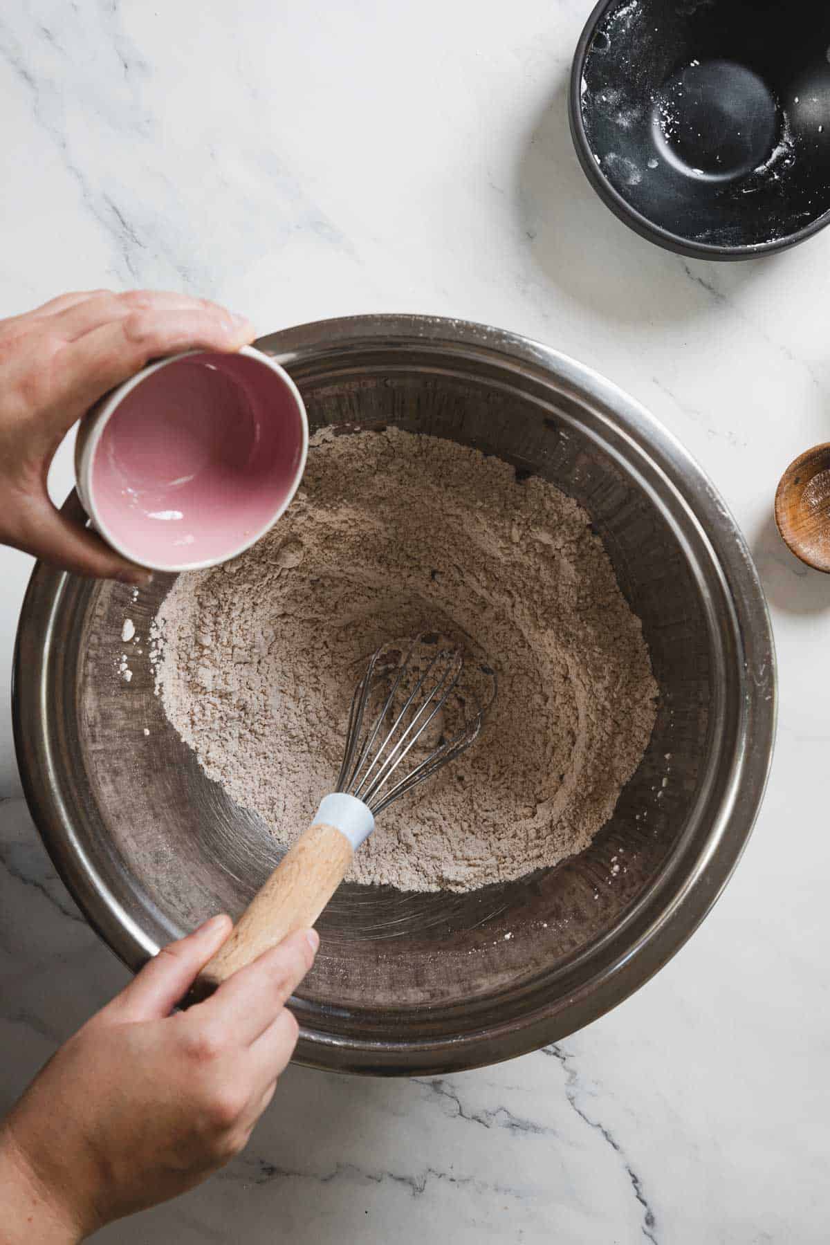 A person is holding a small pink bowl, pouring its contents into a large metal mixing bowl filled with a dry mixture for gingerbread loaf. A whisk is partially submerged in the gluten-free mixture. A black bowl and small wooden bowl are nearby on a white surface.