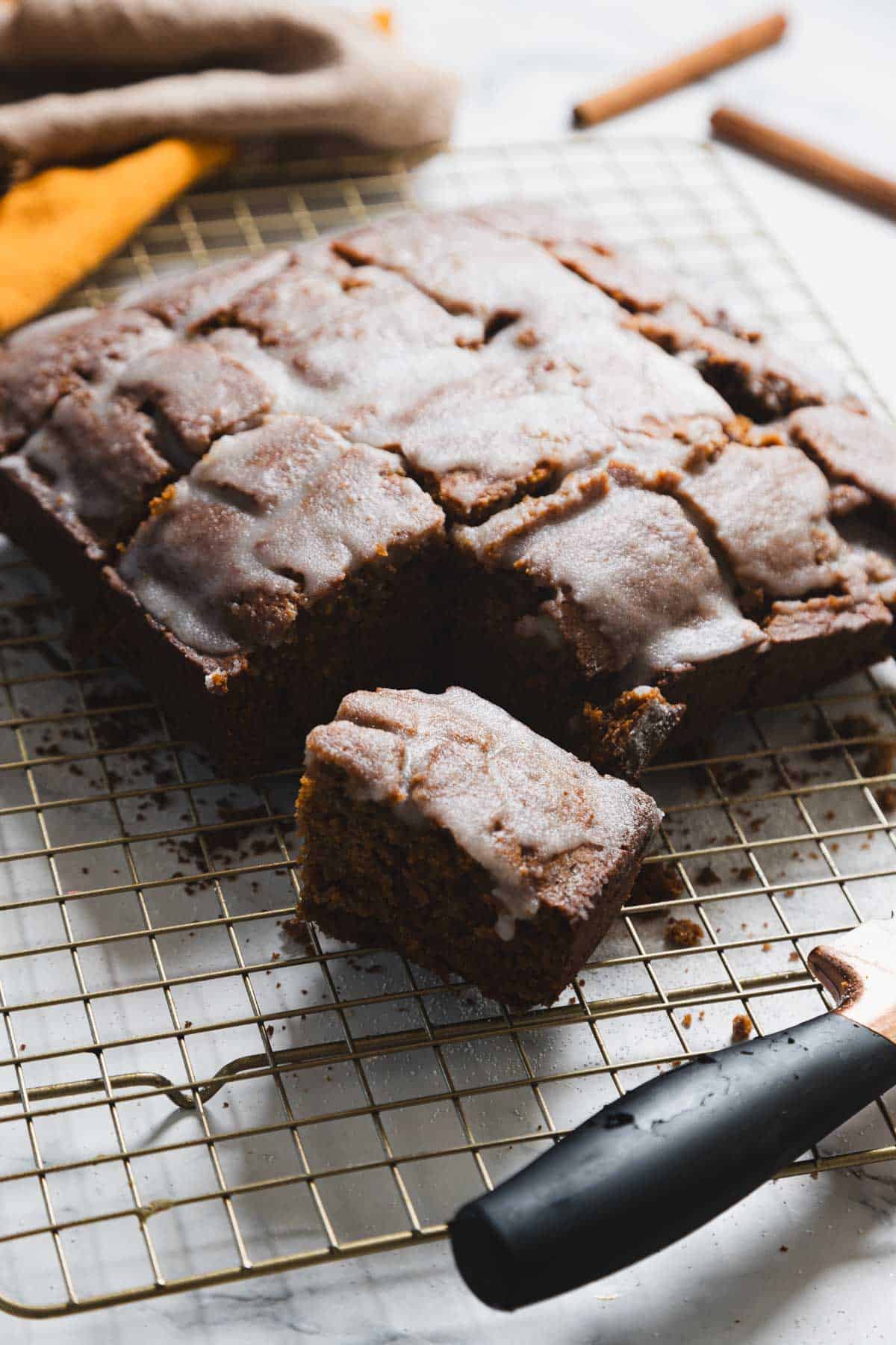 A gluten-free gingerbread loaf with a dusting of glaze sits on a cooling rack. A piece is pulled slightly away, revealing its moist interior. A knife with a black handle rests nearby. Cinnamon sticks and a cloth are visible in the background.