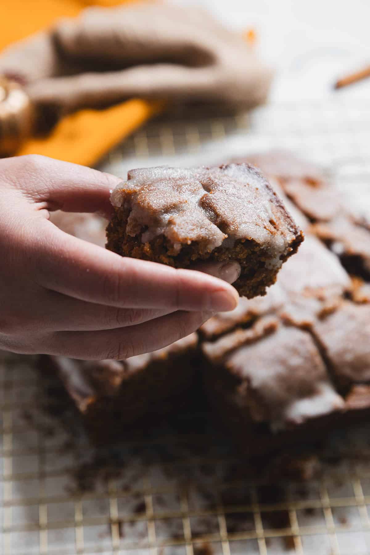 A hand holds a piece of frosted cake with a crumb texture, reminiscent of gingerbread. The cake loaf rests on a cooling rack in the background, with other cut squares visible. A yellow cloth and burlap add warm tones to the scene.