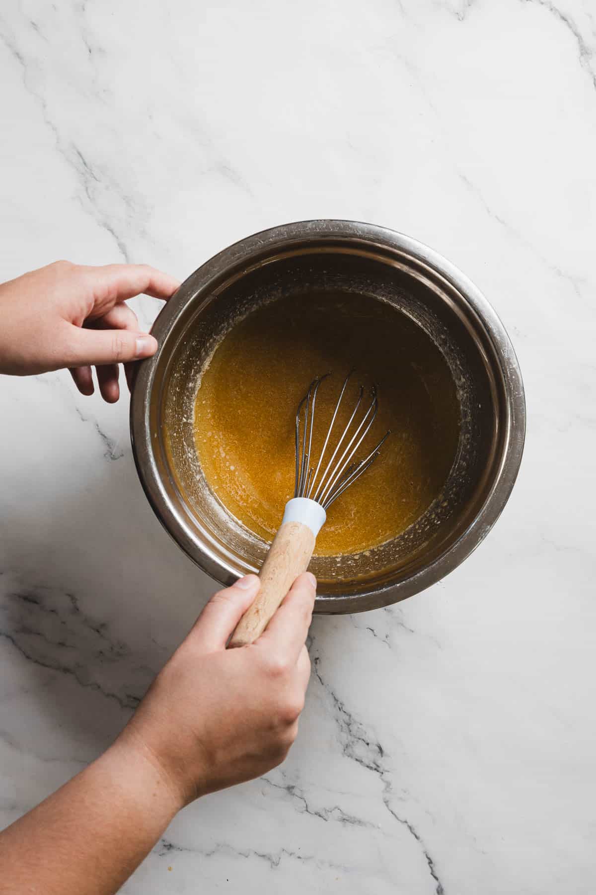 A person holds a metal mixing bowl and stirs a brown gingerbread batter with a wooden-handled whisk. The bowl is placed on a white marble countertop. The image, taken from above, reveals both hands engaged in crafting the perfect gluten-free loaf.