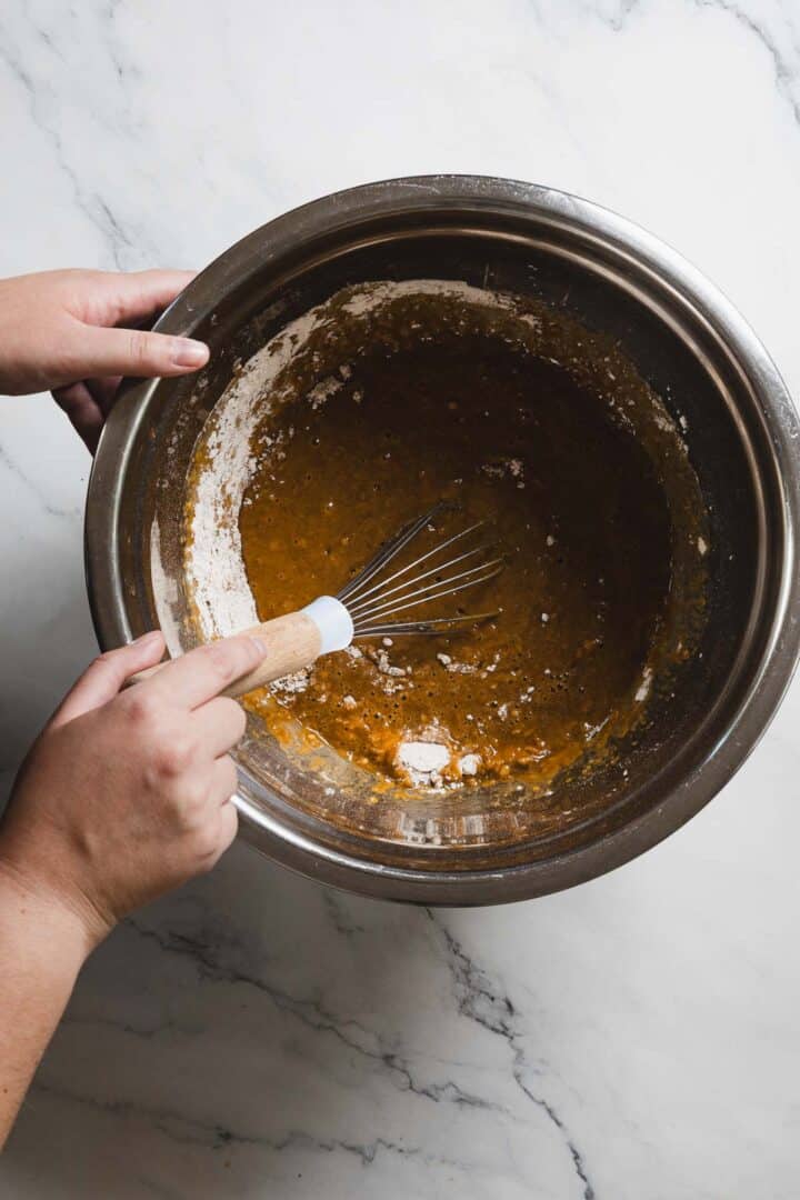 A person mixing a wet brown batter in a stainless steel bowl with a whisk. The gluten-free gingerbread mixture rests on a white marble surface, with some dry flour visible amidst the partially combined ingredients.