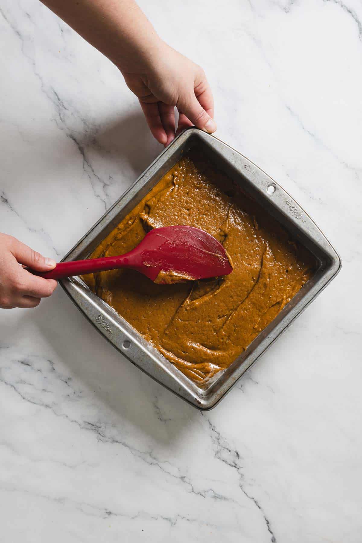 A person is spreading brown gingerbread batter in a square metal baking pan with a red spatula. The pan sits on a white marble surface. This gluten-free mixture appears smooth and evenly distributed.