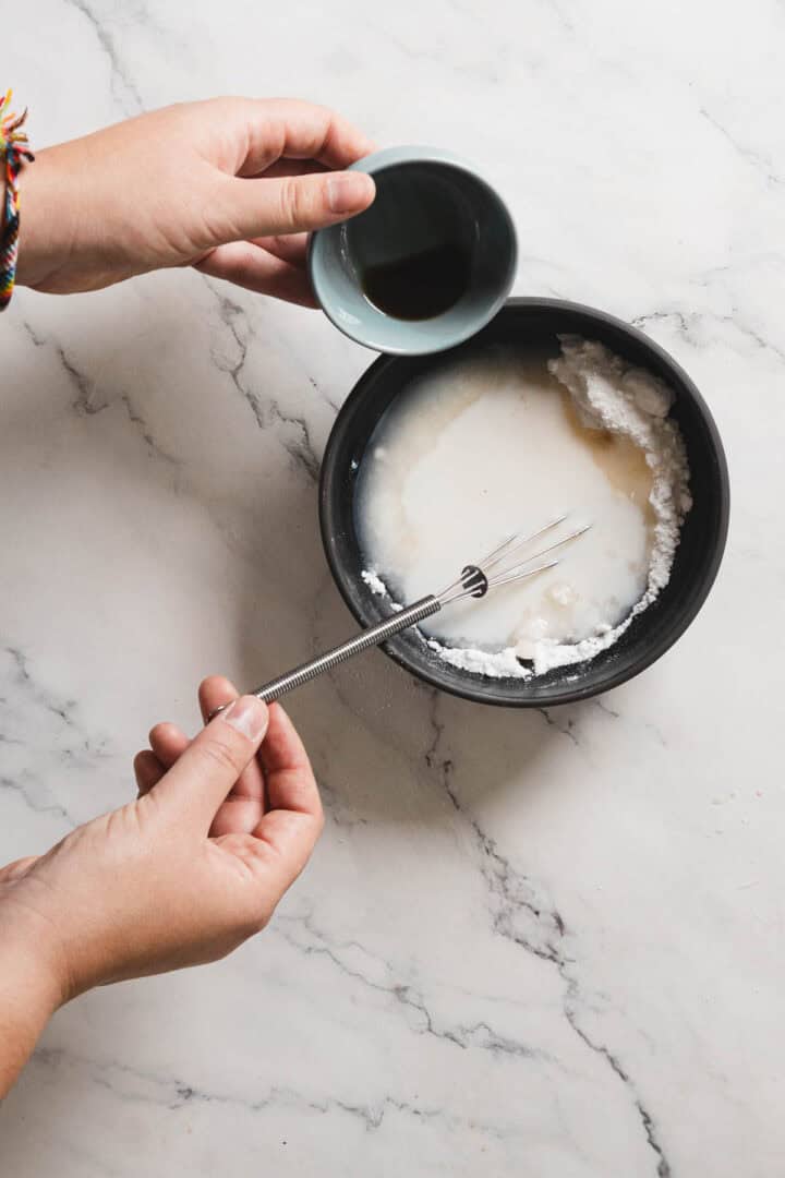 Two hands prepare a mixture in a black bowl on a marble surface. One hand holds a small whisk, while the other pours dark liquid into the larger bowl containing white powder and liquid, crafting the perfect gingerbread blend for an aromatic, gluten-free loaf.