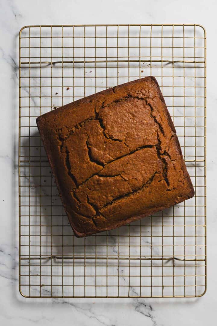 A square, golden-brown gingerbread cake with a cracked top rests on a wire cooling rack. The rack is elegantly placed on a white marble surface, offering a delightful sight.