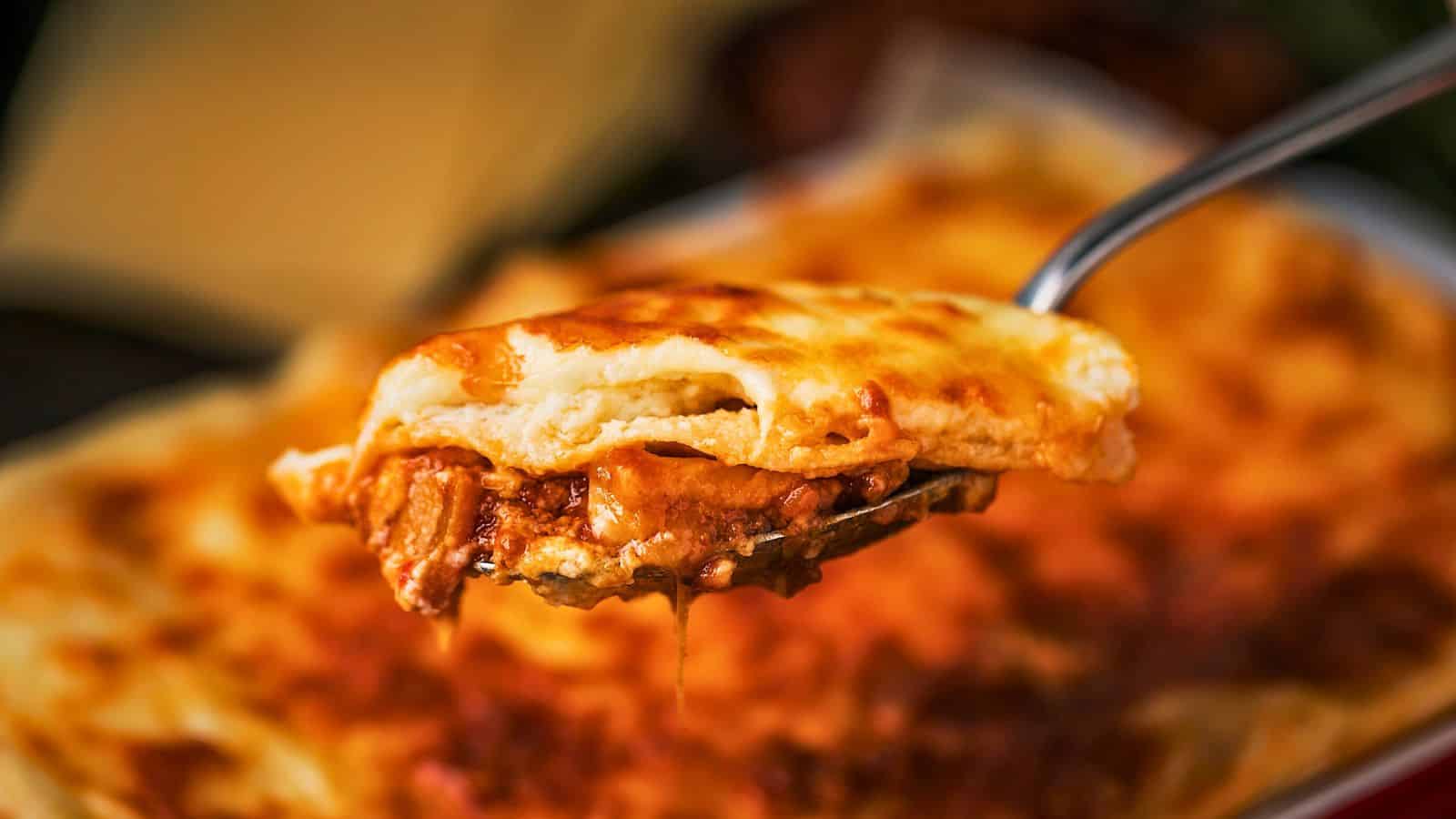 A close-up of a fork holding a piece of lasagna, showing layers of pasta, melted cheese, and visible meat sauce. The background features the rest of the lasagna in a dish, slightly out of focus.