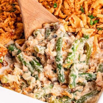 A casserole dish filled with green bean casserole, topped with crispy fried onions. A wooden spoon is scooping a portion, revealing creamy green beans mixed with a thick sauce. A blue and white napkin is partially visible beside the dish.