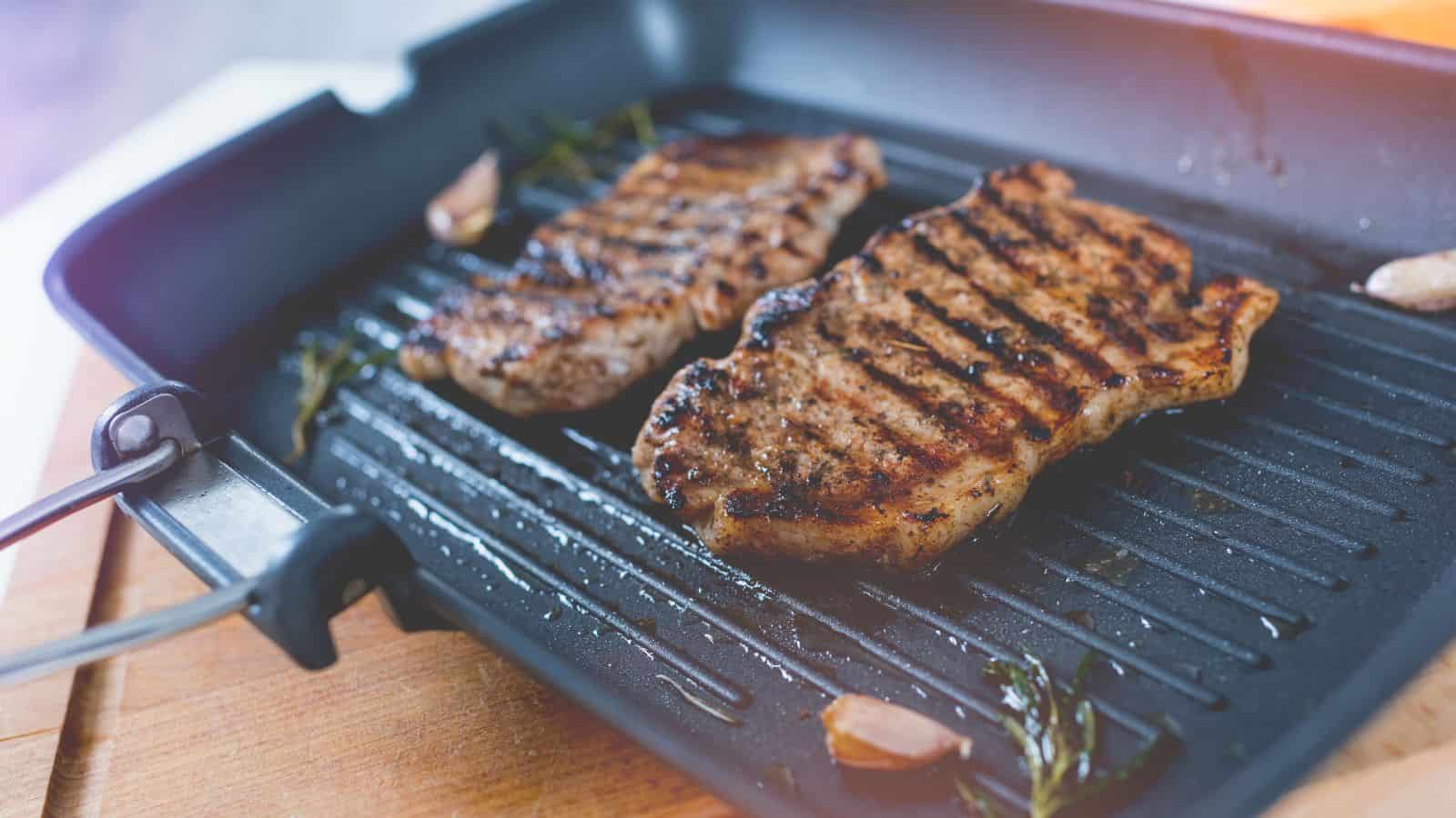 Two grilled steaks with visible grill marks are cooking on a black grill pan. Garlic cloves and rosemary sprigs are scattered around. The pan sits on a wooden cutting board, and soft lighting highlights the scene.