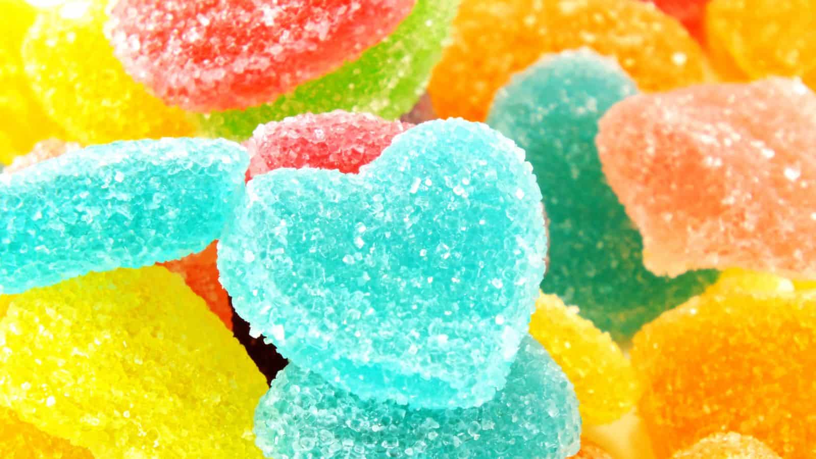 A close-up of colorful sugar-coated gummy candies, including heart-shaped blue gummies, and various oval and circular shapes in red, green, yellow, and orange hues.