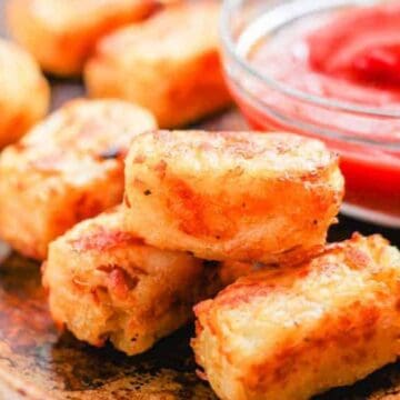 A close-up of a wooden tray showcases crispy, golden-brown tater tots stacked in the foreground, perfect homemade snacks for kids. In the background, a small glass bowl brims with vibrant red ketchup.