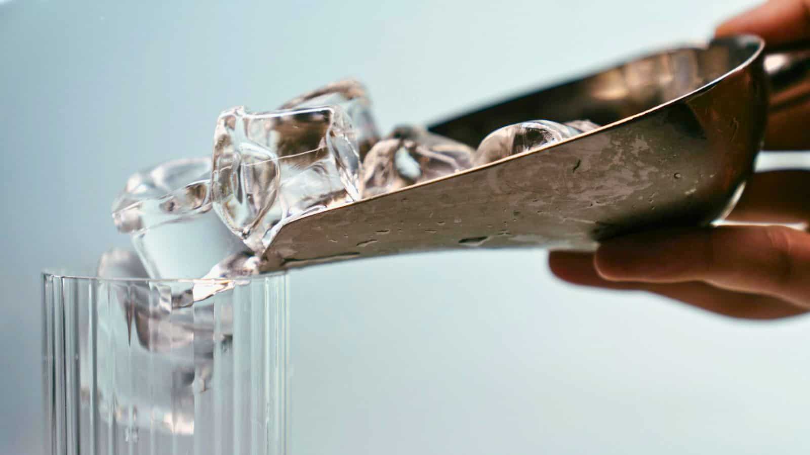 A hand holds a metal scoop, pouring clear ice cubes into a transparent glass. The background is softly blurred, focusing attention on the ice cubes and glass. The scene captures a moment of drink preparation.