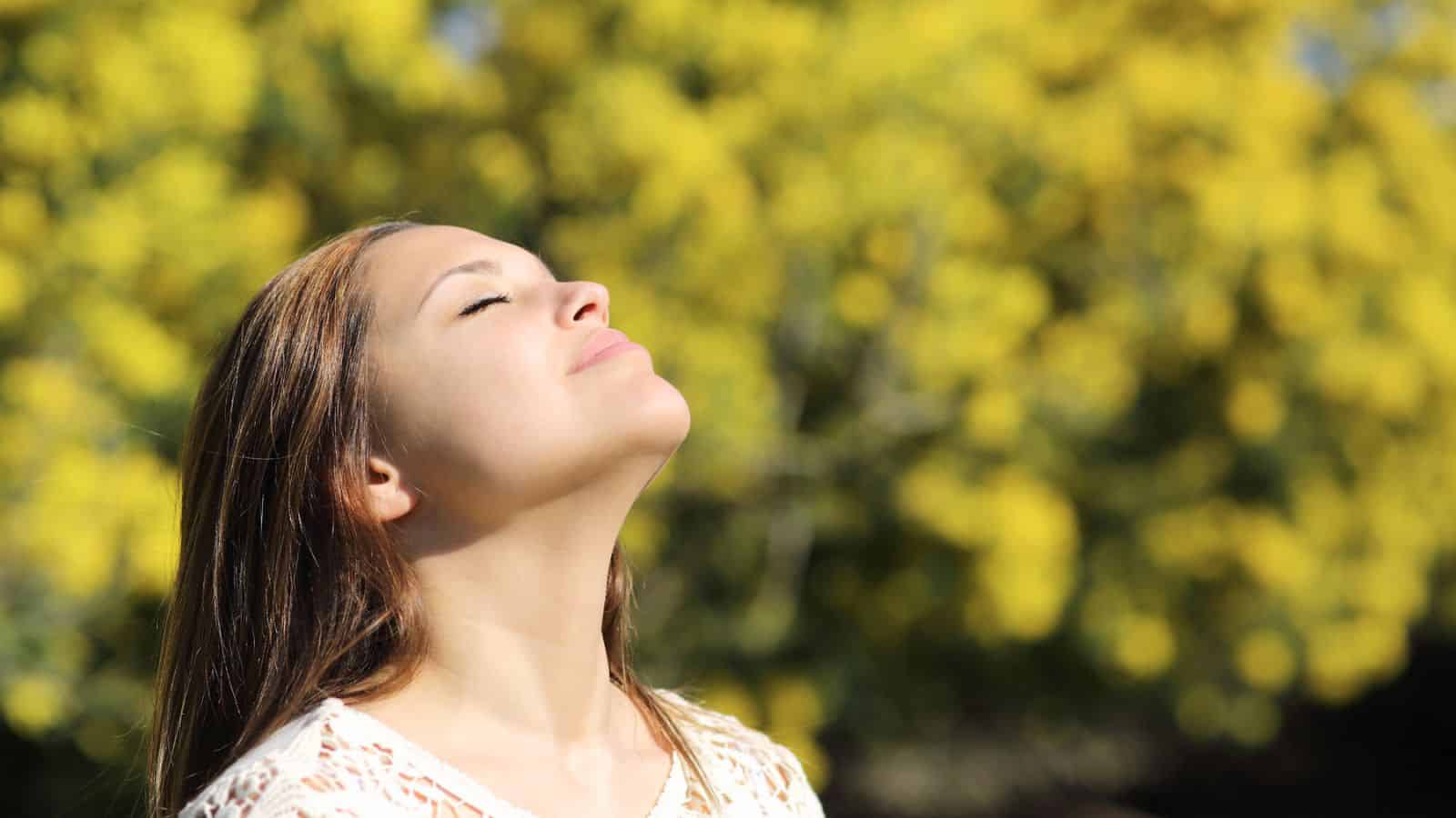 A woman with long hair stands outside, facing the bright sunlight with her eyes closed and a peaceful expression. She is wearing a white top. The background features out-of-focus yellow flowers, suggesting a garden or nature setting.