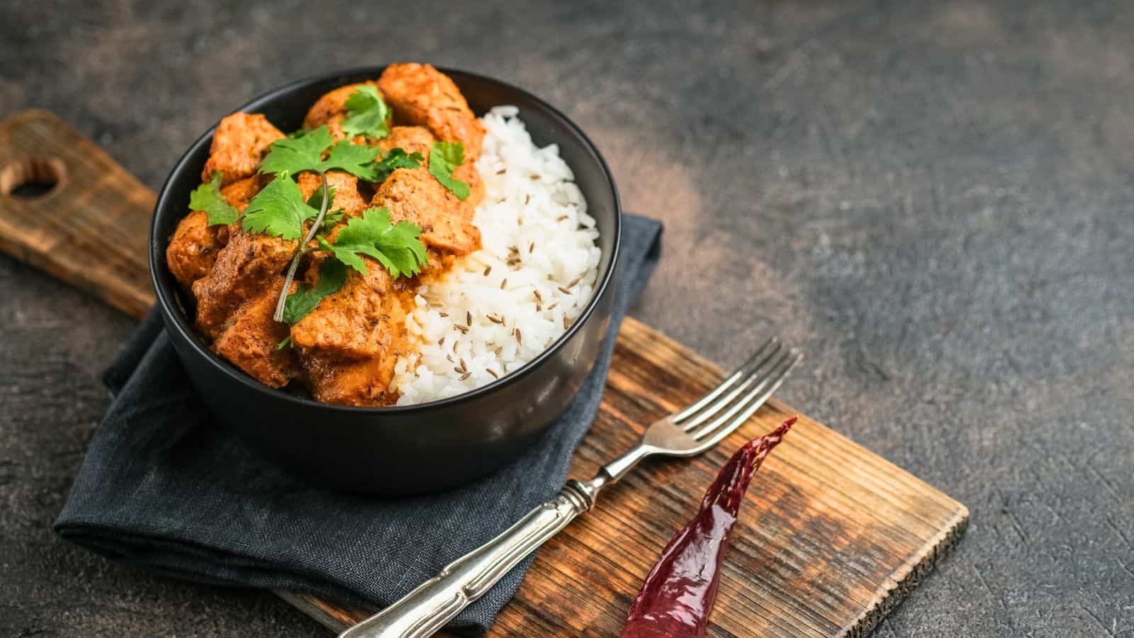 A bowl of rice topped with a curry garnished with cilantro is placed on a wooden board. A fork is beside the bowl on a dark cloth, and a dried chili pepper is positioned near the fork. The background is a textured dark surface.