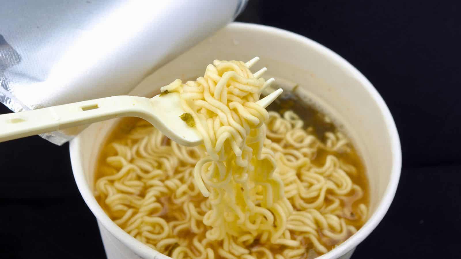 A cup of instant noodles with a plastic fork lifting a portion of cooked noodles. The noodles are in a light broth, and the cup's lid is partially visible, peeled back. The background is black.