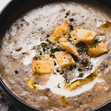 A bowl of creamy mushroom soup topped with croutons, a drizzle of olive oil, and a sprinkle of herbs is placed on a white surface. A sprig of thyme is next to the bowl.