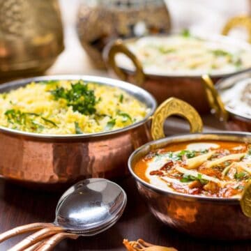 A table with three copper bowls of Indian food. The front dish appears to be curry topped with cream and herbs, the middle dish looks like rice garnished with greens, and the back dish seems to be another curry. Two spoons are placed nearby.