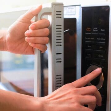 A person adjusts the settings of a microwave oven using a dial while holding the handle with the other hand. The microwave's display and control panel are visible. The person wears a gold bracelet on their left wrist.