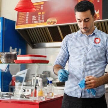 A man in a checkered shirt and apron stands in a fast-food restaurant kitchen preparing food. He wears blue gloves and is near a counter with condiments, a sanitizer bottle, and trays. Red pendant lights hang above, and a refrigerator is in the background.