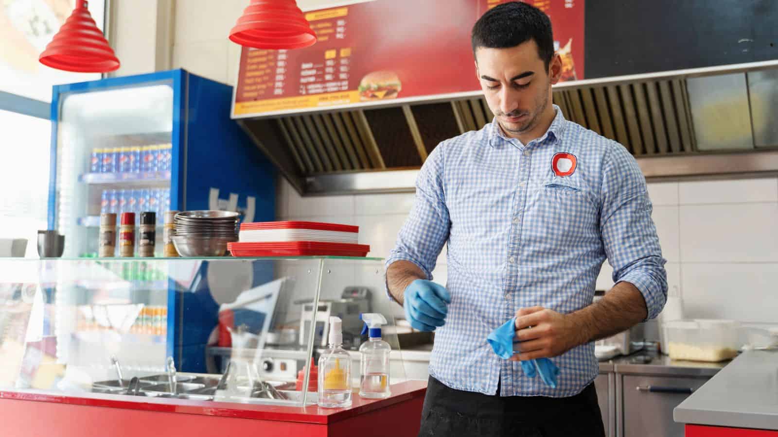 A man in a checkered shirt and apron stands in a fast-food restaurant kitchen preparing food. He wears blue gloves and is near a counter with condiments, a sanitizer bottle, and trays. Red pendant lights hang above, and a refrigerator is in the background.