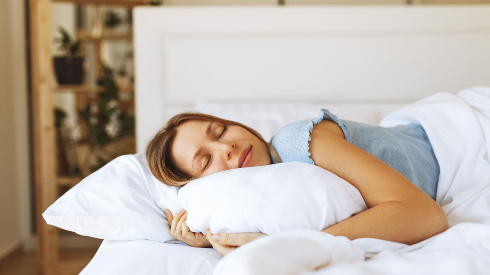 A person with long hair is sleeping peacefully on a white bed, resting their head on a pillow. They are wearing a blue shirt. In the background, there is a wooden shelf with potted plants. The room appears bright and calm.