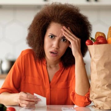 A person in an orange shirt sits at a kitchen counter with a concerned expression, holding a receipt. Beside them is a paper bag filled with groceries, including lettuce, tomatoes, and bread. Various other vegetables are scattered around.