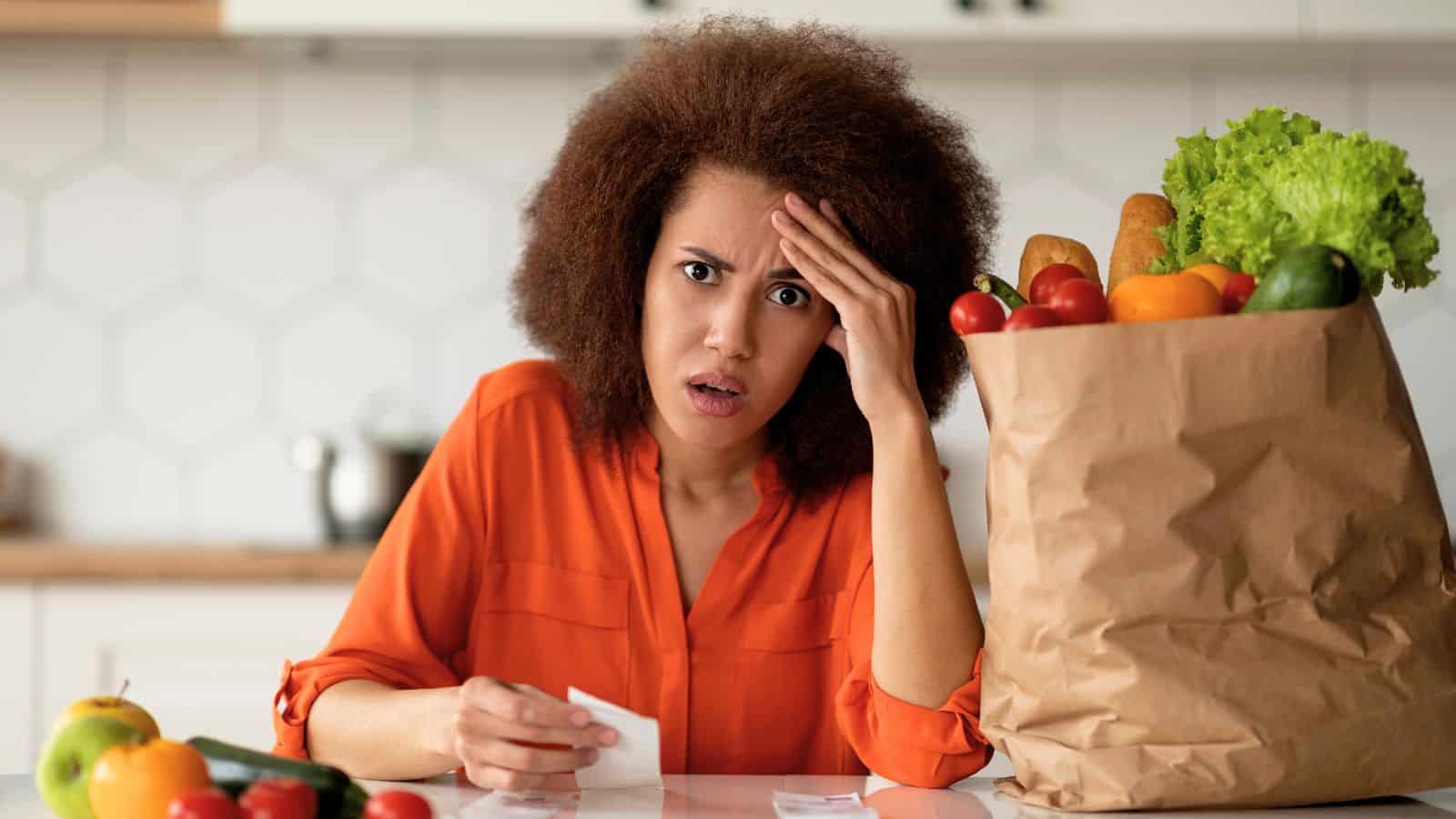 A person in an orange shirt sits at a kitchen counter with a concerned expression, holding a receipt. Beside them is a paper bag filled with groceries, including lettuce, tomatoes, and bread. Various other vegetables are scattered around.