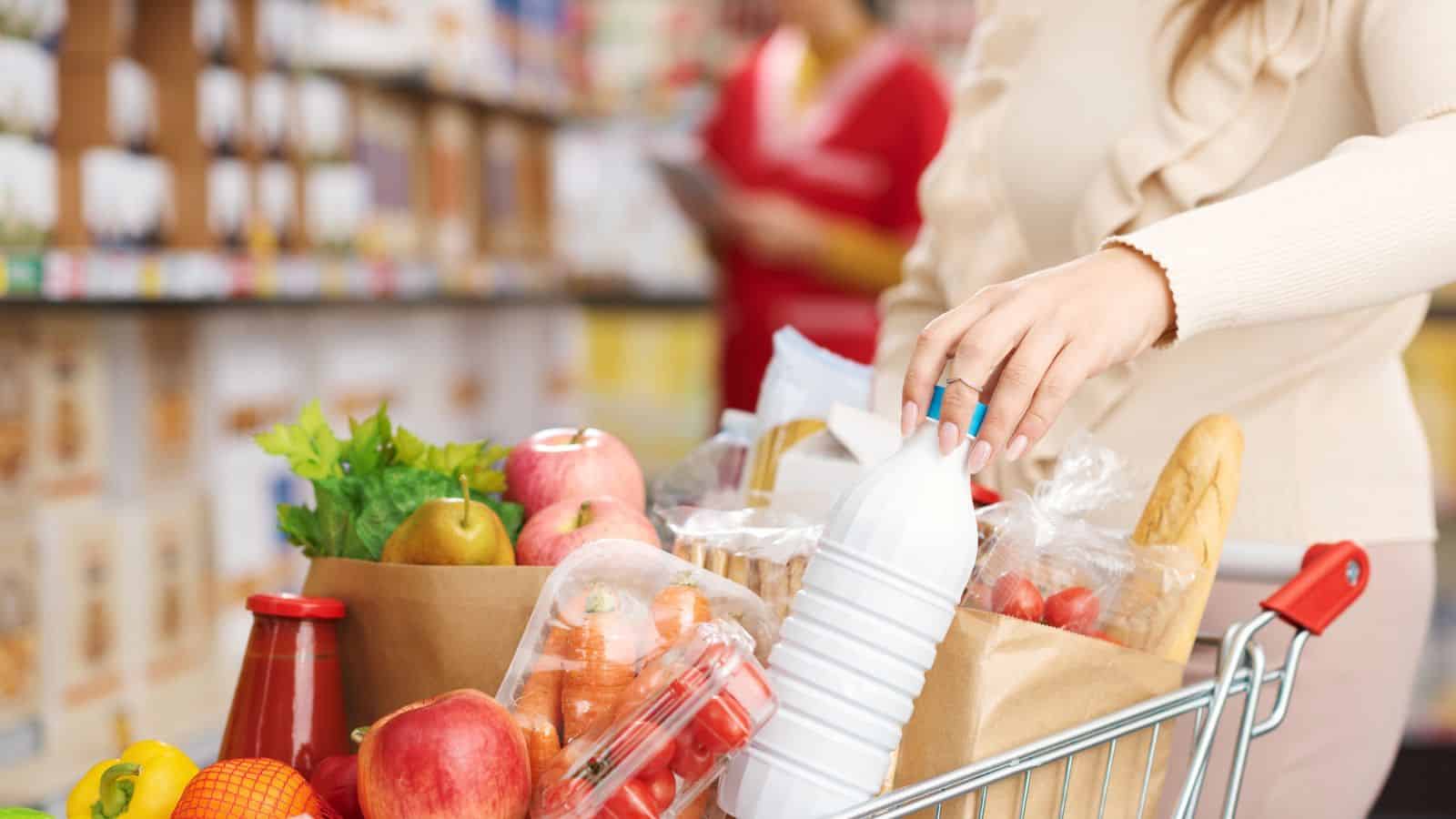 A person with long hair reaches for a bottle in a shopping cart filled with groceries, including apples, lettuce, cherries, and a baguette. Shelves and another person in red uniform are visible in the blurred background.