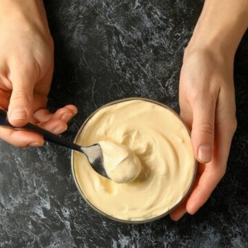 Two hands hold a glass bowl filled with creamy mayonnaise. A black spoon is scooping some mayonnaise from the bowl. The background is a dark, textured surface.