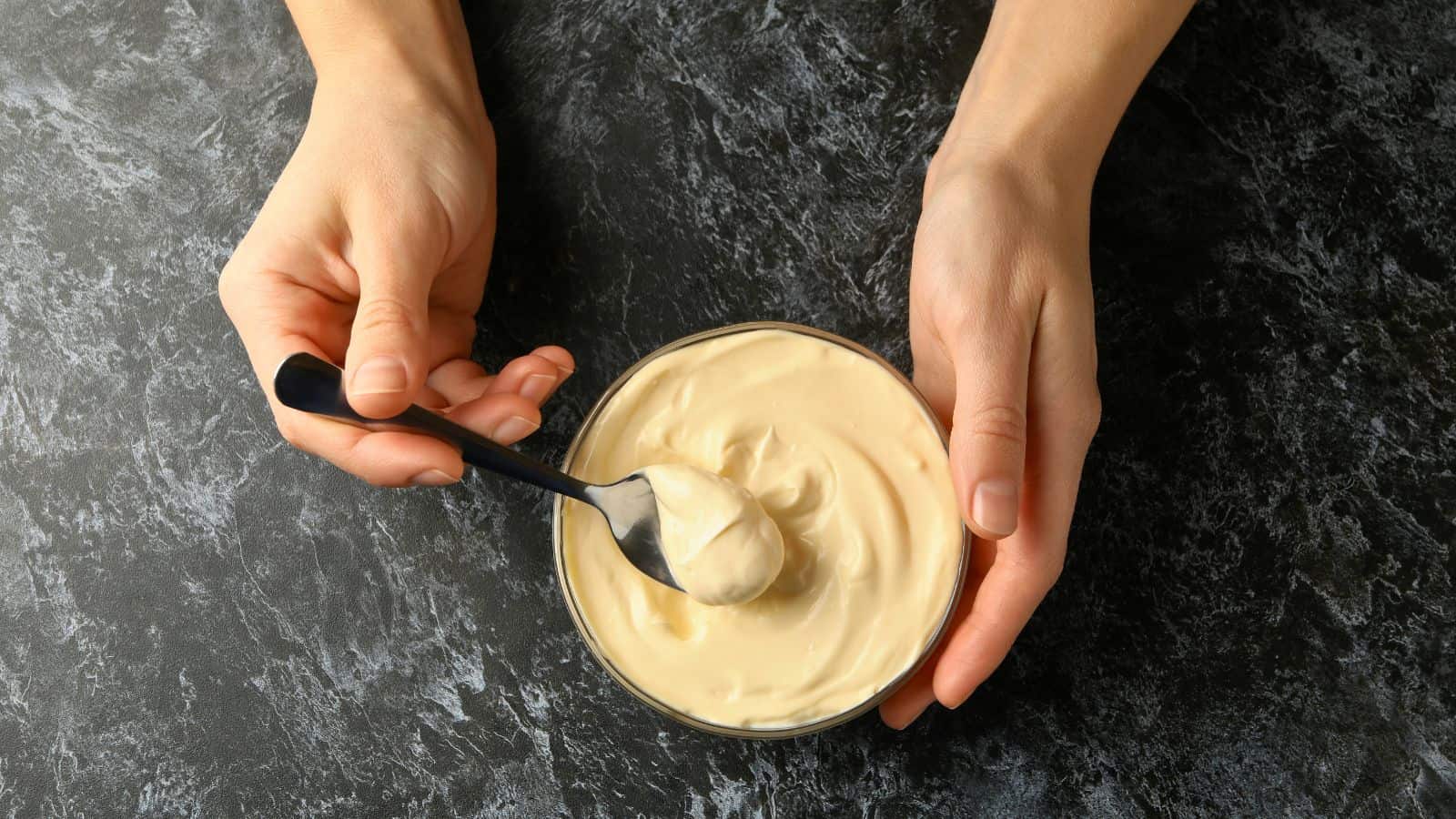 Two hands hold a glass bowl filled with creamy mayonnaise. A black spoon is scooping some mayonnaise from the bowl. The background is a dark, textured surface.