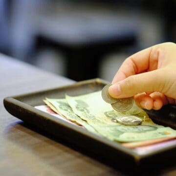 A hand places coins onto a tray with paper currency on a wooden table.