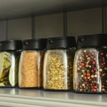 Four glass spice jars with black lids are lined up on a shelf. The jars contain bay leaves, brown sugar, mixed herbs, and whole peppercorns. White bowls are visible on either side of the jars.