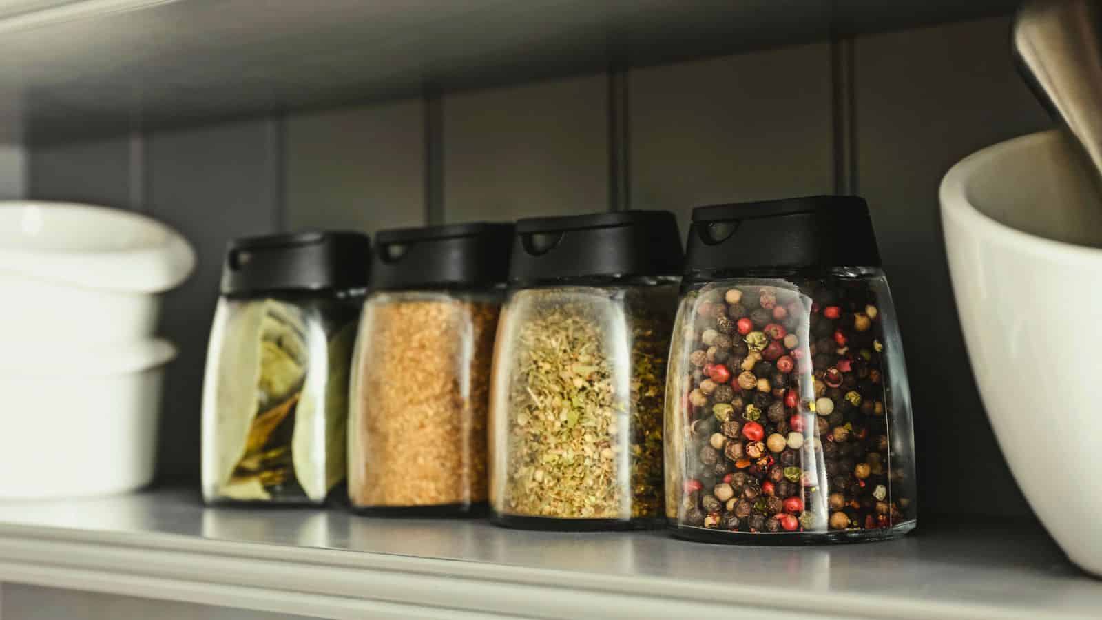 Four glass spice jars with black lids are lined up on a shelf. The jars contain bay leaves, brown sugar, mixed herbs, and whole peppercorns. White bowls are visible on either side of the jars.