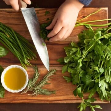 A person chops green herbs on a wooden cutting board. There are bunches of basil, chives, rosemary, and parsley. A small white bowl filled with yellow olive oil is also on the board.